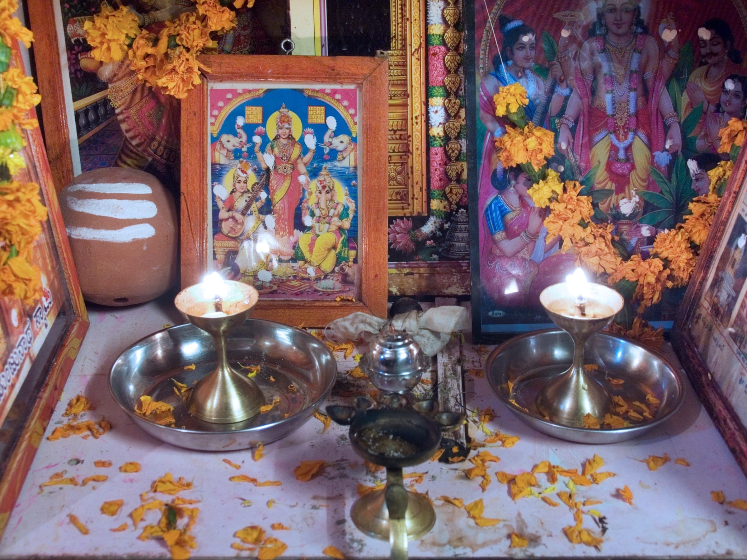 A family altar in an Indian home, deity prints framed with marigolds, brass aarti lamps lit, flower petals scattered on the threshold