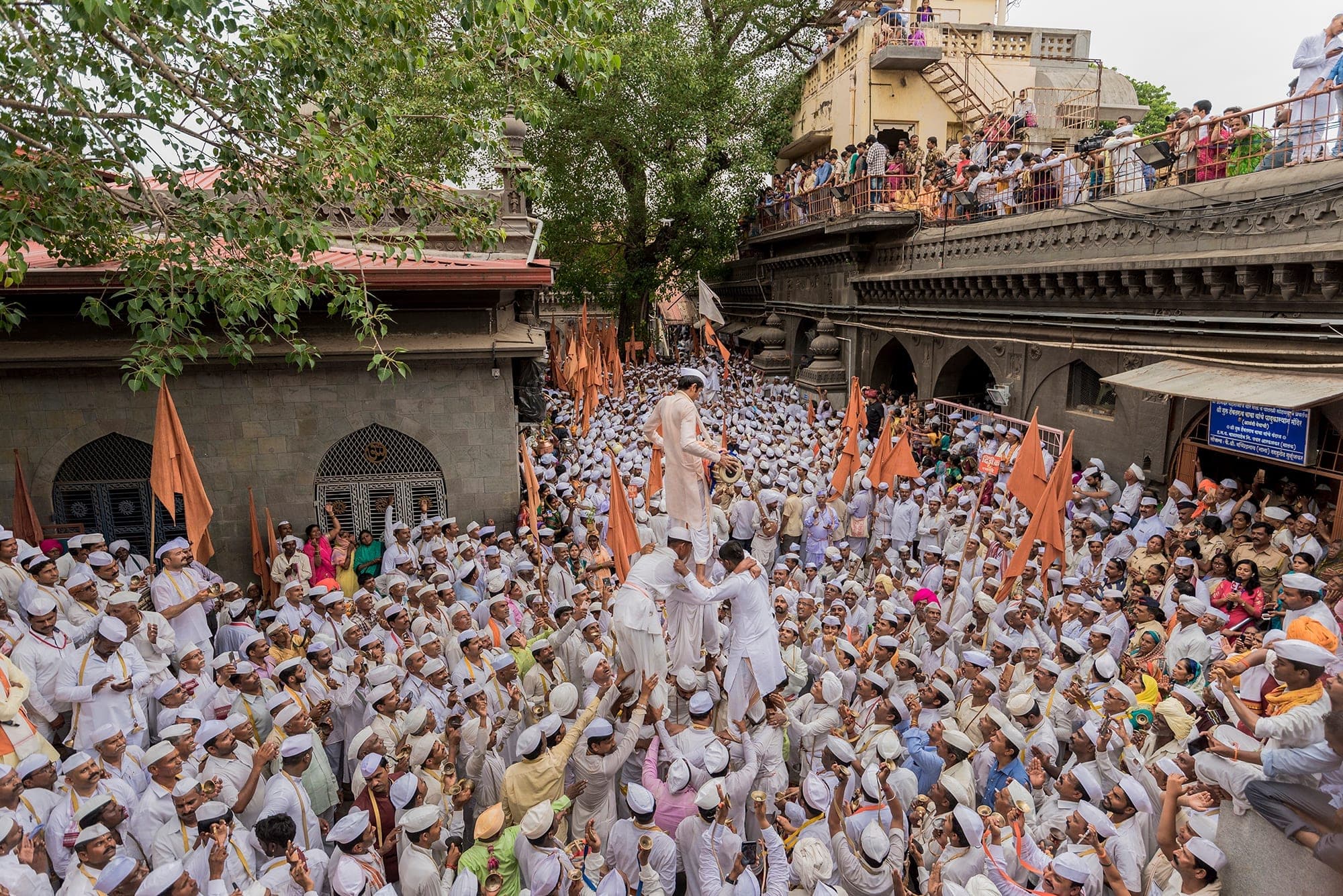 A sea of warkari devotees in white caps streaming up to a stone temple gateway, saffron flags raised